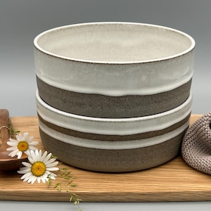 Puede incluir: Dos cuencos de cer&aacute;mica apilados, con un dise&ntilde;o de dos tonos: parte superior blanca y una banda marr&oacute;n. Los cuencos est&aacute;n sobre una tabla de madera con una cuchara de madera y margaritas. Tambi&eacute;n hay un pa&ntilde;o de punto beige.