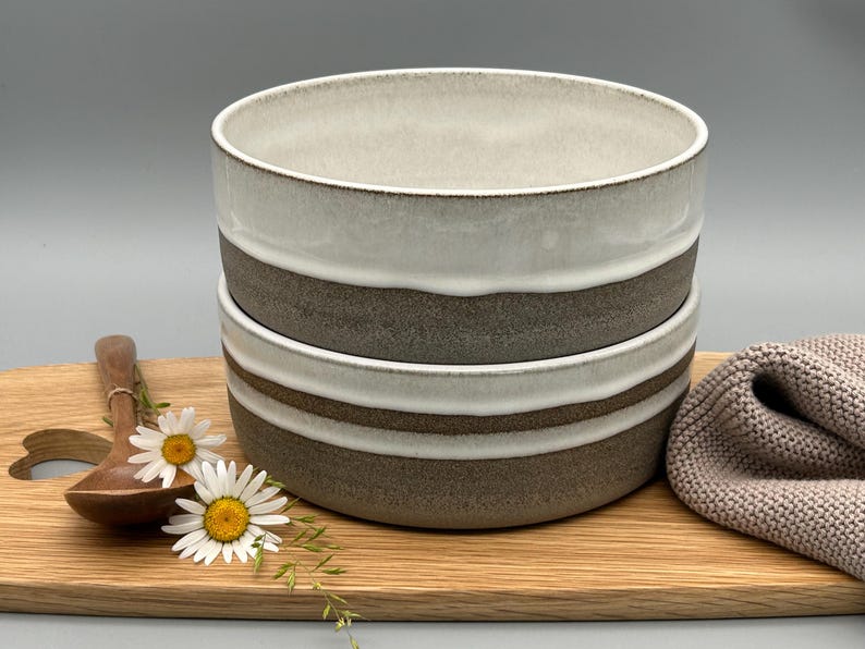Puede incluir: Dos cuencos de cer&aacute;mica apilados, con un dise&ntilde;o de dos tonos: parte superior blanca y una banda marr&oacute;n. Los cuencos est&aacute;n sobre una tabla de madera con una cuchara de madera y margaritas. Tambi&eacute;n hay un pa&ntilde;o de punto beige.