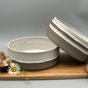 Puede incluir: Dos cuencos de cer&aacute;mica en tonos blancos y marrones, colocados sobre una tabla de cortar de madera. Un cuenco est&aacute; en posici&oacute;n vertical, el otro est&aacute; inclinado. Una cuchara de madera y margaritas tambi&eacute;n est&aacute;n en la tabla. Un art&iacute;culo de punto beige est&aacute; a la derecha.