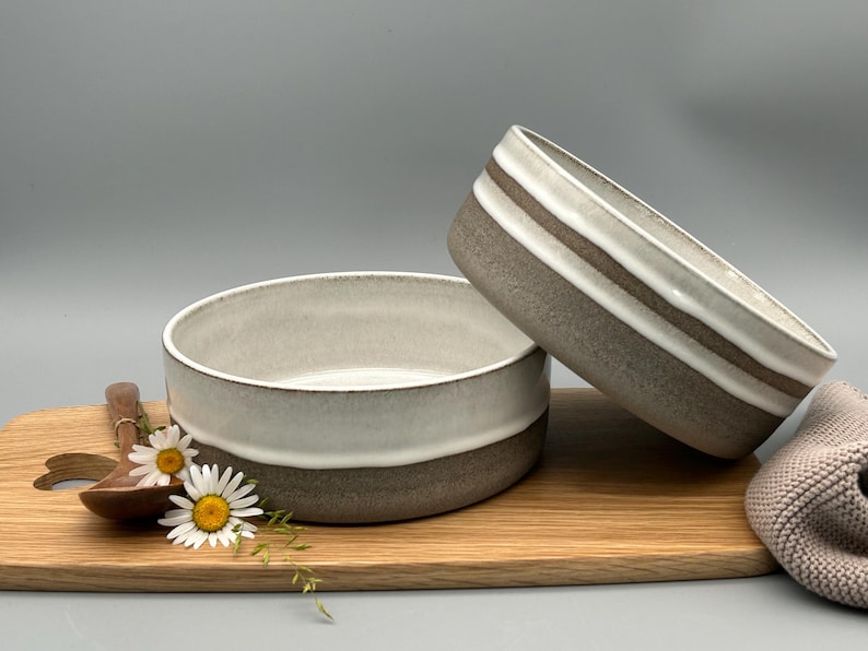 Puede incluir: Dos cuencos de cer&aacute;mica en tonos blancos y marrones, colocados sobre una tabla de cortar de madera. Un cuenco est&aacute; en posici&oacute;n vertical, el otro est&aacute; inclinado. Una cuchara de madera y margaritas tambi&eacute;n est&aacute;n en la tabla. Un art&iacute;culo de punto beige est&aacute; a la derecha.