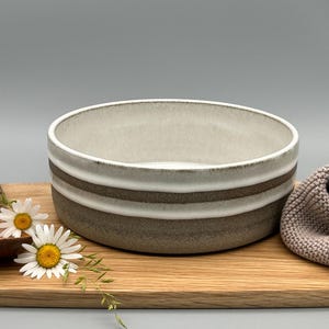 Puede incluir: Un cuenco de cer&aacute;mica con interior beige claro y exterior a rayas blancas y marrones sobre una tabla de cortar de madera. Una cuchara de madera y dos margaritas est&aacute;n junto al cuenco. Un pa&ntilde;o de punto beige est&aacute; a la derecha.