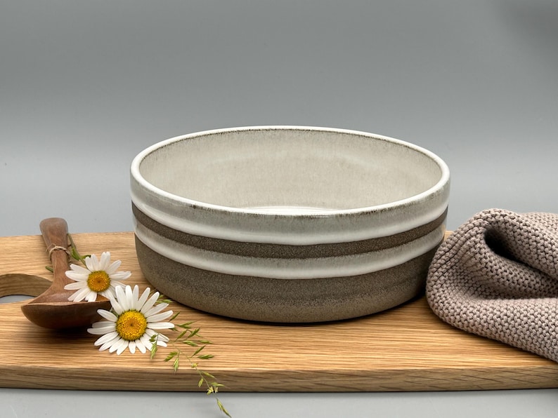 Puede incluir: Un cuenco de cer&aacute;mica con interior beige claro y exterior a rayas blancas y marrones sobre una tabla de cortar de madera. Una cuchara de madera y dos margaritas est&aacute;n junto al cuenco. Un pa&ntilde;o de punto beige est&aacute; a la derecha.