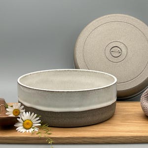 Puede incluir: Un cuenco de cer&aacute;mica con tapa, con un dise&ntilde;o bicolor: blanco arriba, marr&oacute;n abajo. La tapa refleja el dise&ntilde;o. El cuenco est&aacute; sobre una tabla de madera, con una cuchara de madera y flores. Medidas en cent&iacute;metros.