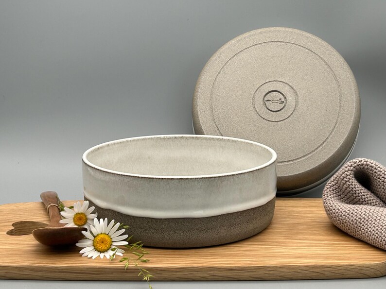 Puede incluir: Un cuenco de cer&aacute;mica con tapa, con un dise&ntilde;o bicolor: blanco arriba, marr&oacute;n abajo. La tapa refleja el dise&ntilde;o. El cuenco est&aacute; sobre una tabla de madera, con una cuchara de madera y flores. Medidas en cent&iacute;metros.