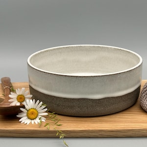 Puede incluir: Cuenco de cer&aacute;mica de dos tonos, gris claro en la parte superior y marr&oacute;n oscuro en la inferior, sobre una tabla de madera. Una cuchara de madera y margaritas junto al cuenco. Un art&iacute;culo de punto gris a la derecha.