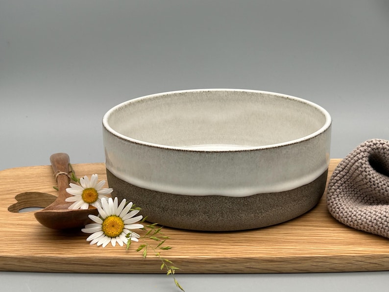 Puede incluir: Cuenco de cer&aacute;mica de dos tonos, gris claro en la parte superior y marr&oacute;n oscuro en la inferior, sobre una tabla de madera. Una cuchara de madera y margaritas junto al cuenco. Un art&iacute;culo de punto gris a la derecha.