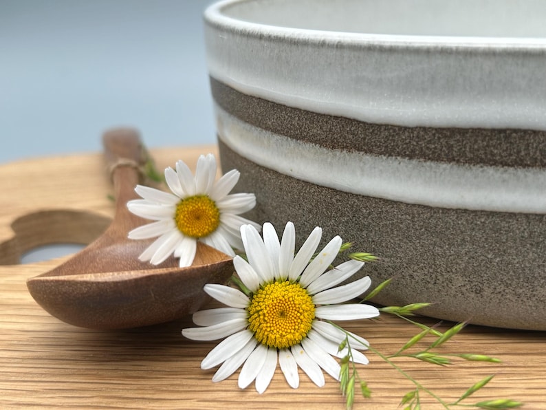 Puede incluir: Un cuenco de cer&aacute;mica con un dise&ntilde;o a rayas blancas y marrones, una cuchara de madera y dos margaritas con p&eacute;talos blancos y centros amarillos. Los art&iacute;culos est&aacute;n dispuestos sobre una superficie de madera.