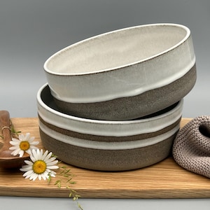 Puede incluir: Dos cuencos de cer&aacute;mica apilados, blancos y grises con una banda marr&oacute;n, sobre una tabla de madera. Tambi&eacute;n hay una cuchara de madera, margaritas y un pa&ntilde;o beige. Los cuencos son ideales para servir sopa o cereales.