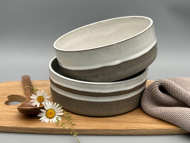 Puede incluir: Dos cuencos de cer&aacute;mica apilados, blancos y grises con una banda marr&oacute;n, sobre una tabla de madera. Tambi&eacute;n hay una cuchara de madera, margaritas y un pa&ntilde;o beige. Los cuencos son ideales para servir sopa o cereales.
