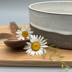 Puede incluir: Un cuenco de cer&aacute;mica con esmalte blanco y gris, una cuchara de madera y una tabla de cortar de madera. Dos margaritas y hierba se colocan en la tabla. El cuenco tiene unos 15 cm de di&aacute;metro.