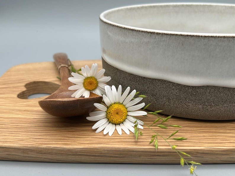 Puede incluir: Un cuenco de cer&aacute;mica con esmalte blanco y gris, una cuchara de madera y una tabla de cortar de madera. Dos margaritas y hierba se colocan en la tabla. El cuenco tiene unos 15 cm de di&aacute;metro.