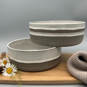 Puede incluir: Dos cuencos de cer&aacute;mica con un dise&ntilde;o en capas. Los cuencos tienen una parte superior blanca, una franja marr&oacute;n y una base gris. Los cuencos est&aacute;n sobre una tabla de cortar de madera con una cuchara de madera y margaritas.
