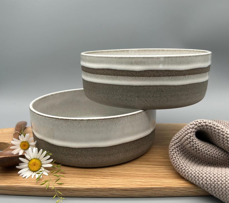 Puede incluir: Dos cuencos de cer&aacute;mica con un dise&ntilde;o en capas. Los cuencos tienen una parte superior blanca, una franja marr&oacute;n y una base gris. Los cuencos est&aacute;n sobre una tabla de cortar de madera con una cuchara de madera y margaritas.