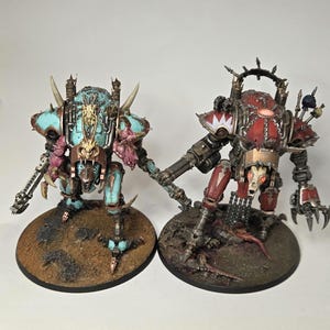 May include: Two miniature robot figures on circular bases. The left figure is turquoise and metallic with bone-like details. The right figure is red and metallic with a spiked crown, chains, and a skull-like face. Both have mechanical limbs.