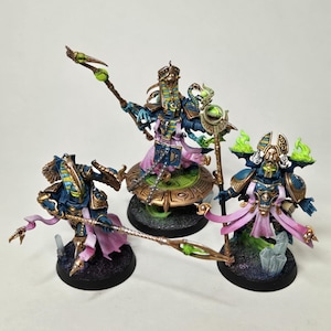 Puede incluir: Tres figuras en miniatura pintadas de azul y dorado con detalles verdes y rosas. Las figuras llevan túnicas elaboradas y sostienen bastones. Las figuras están de pie sobre bases circulares.