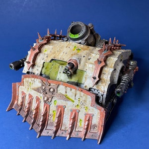 May include: A white and green model of a heavily armored vehicle with a large, spiked blade on the front. The vehicle has a large, round cannon on top and several smaller cannons on the sides. The model is painted in a weathered, battle-damaged style.