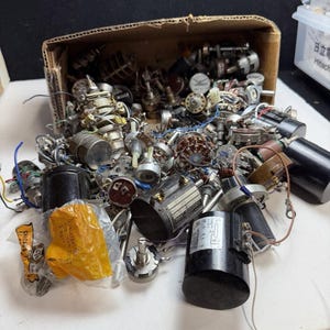 May include: A collection of various electronic components, including multiple potentiometers, rotary switches, and cylindrical capacitors, some with attached wires, spilling out of a brown cardboard box onto a white surface.