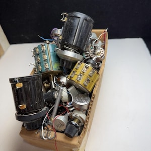 May include: A cardboard box filled with an assortment of electronic components, including various rotary potentiometers, switches, and dials in black, teal, and yellow colors, featuring metallic shafts and attached thin colored electrical wires.