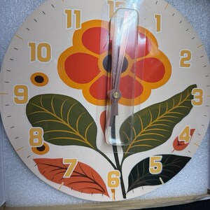 May include: A round wall clock with a cream-colored face featuring a large orange and yellow flower graphic with green leaves. The clock has yellow numbers from 1 to 12 arranged around the perimeter and thin, dark-colored hands protected by a clear plastic cover.