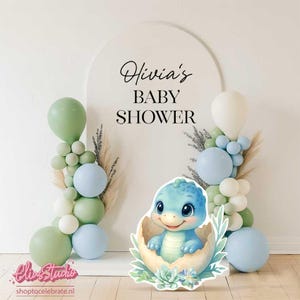 May include: A baby shower decoration featuring a blue dinosaur hatching from an egg, surrounded by pastel balloons and floral accents. The arch backdrop reads "Olivia's BABY SHOWER".