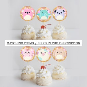 May include: Six cupcake toppers featuring various animal designs. The toppers are round and have a gold glitter background. The animals include a koala, a dragon, a panda, a cat, a unicorn, and a bunny. The toppers are placed on top of cupcakes with white frosting.