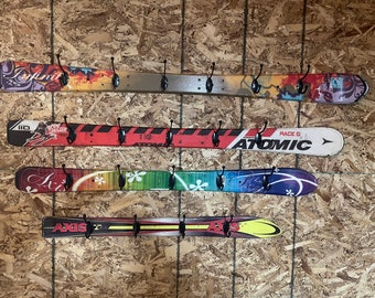 Może przedstawiać: Cztery ścienne wieszaki wykonane z używanych nart. Każda narta posiada odmienną, kolorową grafikę, w tym napisy Infinite, Bode Miller, Atomic oraz Axis. Każda narta jest wyposażona w pięć czarnych metalowych haczyków do wieszania rzeczy.