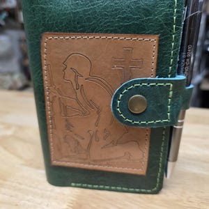 May include: Green leather journal with a brown leather patch featuring an etched image of a soldier kneeling before a cross. The journal has a snap closure and a pen holder. The stitching is a contrasting light green color.