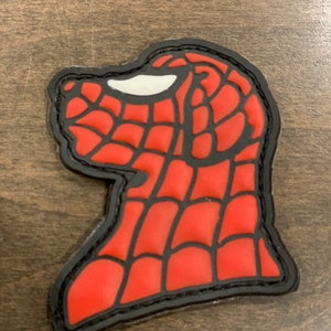 May include: Red and black embroidered patch with a Spiderman head design. The patch has a black outline and a white eye.