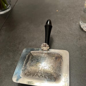 May include: Silver-plated butter dish with a black handle. The dish has a rectangular shape with ornate floral and scrollwork detailing on the lid. A small shell-shaped accent is at the base of the handle.