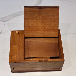 May include: A rectangular wooden box with a hinged lid that is propped open. The box is constructed from medium-brown wood with a visible grain. A small, light-colored wooden knob is positioned on the left side of the top surface of the box.