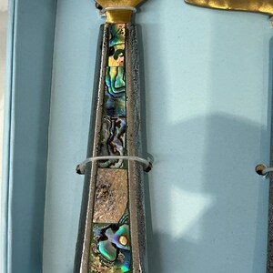 K&ouml;nnte beinhalten: Nahaufnahme eines K&auml;semesser- und Streuset-Sets. Die Griffe sind silberfarben mit schillernden Abalone-Muscheleinlagen. Die Klingen sind goldfarben. Das Set wird in einer hellblauen Box pr&auml;sentiert.