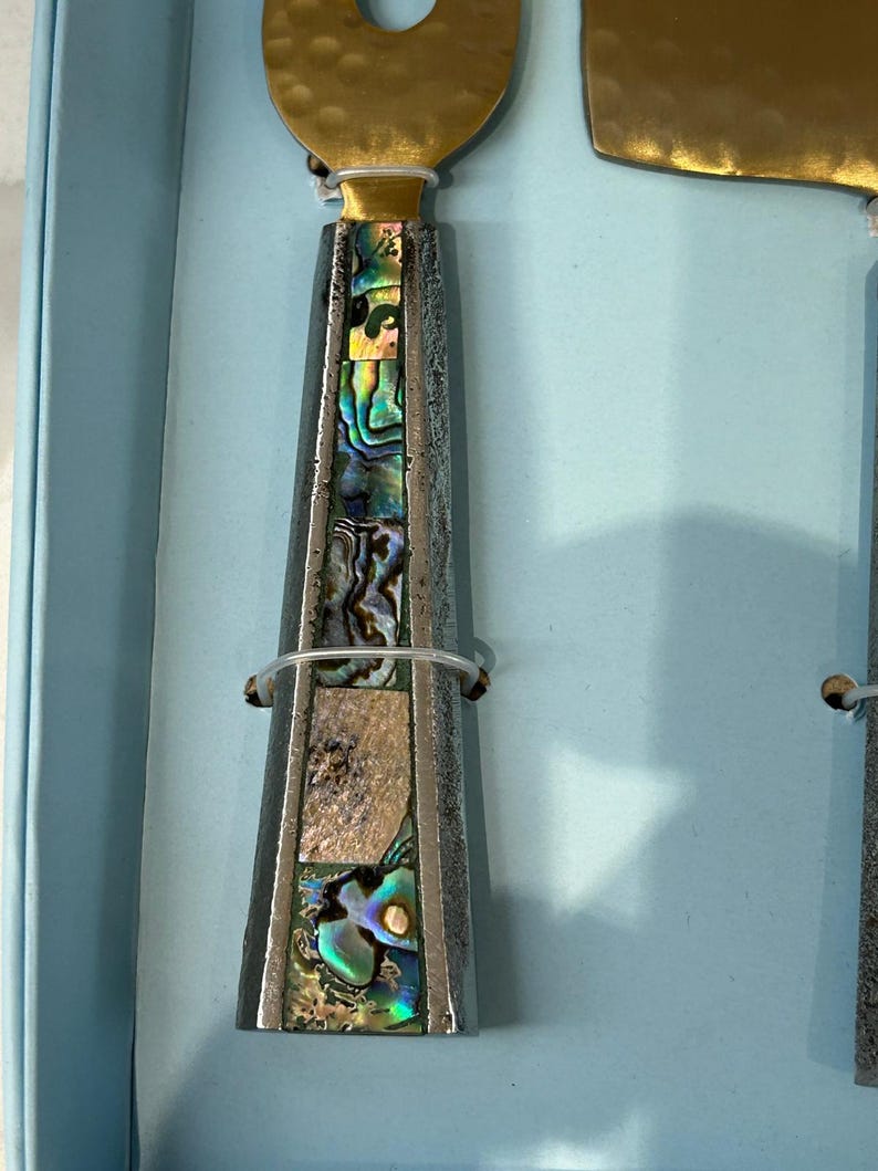 K&ouml;nnte beinhalten: Nahaufnahme eines K&auml;semesser- und Streuset-Sets. Die Griffe sind silberfarben mit schillernden Abalone-Muscheleinlagen. Die Klingen sind goldfarben. Das Set wird in einer hellblauen Box pr&auml;sentiert.