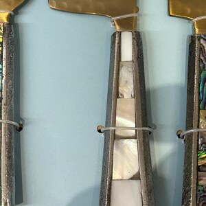 K&ouml;nnte beinhalten: Nahaufnahme von drei K&auml;semessern. Die Klingen sind goldfarben und die Griffe sind silberfarben mit eingelegten Perlmutt- und Abalone-Muschel-Akzenten. Die Messer sind mit d&uuml;nnem Draht zusammengebunden.