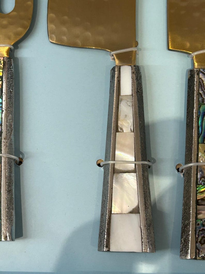 K&ouml;nnte beinhalten: Nahaufnahme von drei K&auml;semessern. Die Klingen sind goldfarben und die Griffe sind silberfarben mit eingelegten Perlmutt- und Abalone-Muschel-Akzenten. Die Messer sind mit d&uuml;nnem Draht zusammengebunden.