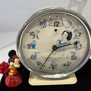 May include: Vintage Disney Snow White alarm clock with a cream face and silver metal frame. The clock features illustrations of Snow White and the Seven Dwarfs. The clock is on a cream-coloured base.