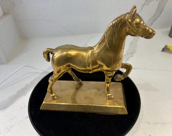 May include: A golden brass horse figurine in a trotting pose. The sculpture is mounted on a rectangular base. The horse has detailed features, including a flowing mane and a harness. The figurine is displayed on a black surface.