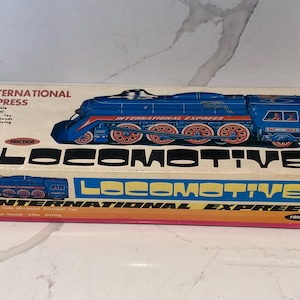 Könnte beinhalten: Ein rechteckiger Pappkarton für eine International Express Reibungsspielzeug-Lokomotive. Der Karton zeigt eine blaue Zugillustration, den Text International Express und das Wort LOCOMOTIVE in großen schwarzen Buchstaben auf weißem Grund.