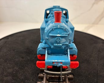 May include: A blue toy steam locomotive engine featuring a bright red chimney stack and red buffer beams at the front. The model train is positioned on a short section of black plastic railway track, showcasing its front-facing design and details.