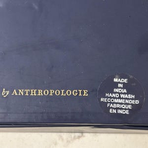 K&ouml;nnte beinhalten: Nahaufnahme einer marineblauen Box mit goldenem Text "by ANTHROPOLOGIE". Ein runder Aufkleber besagt "MADE IN INDIA, HAND WASH RECOMMENDED, FABRIQUE EN INDE."