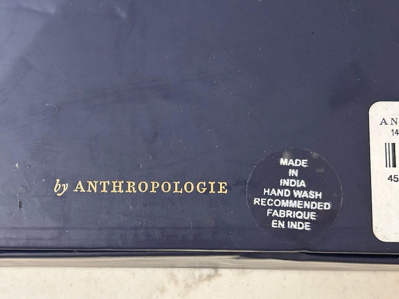 K&ouml;nnte beinhalten: Nahaufnahme einer marineblauen Box mit goldenem Text "by ANTHROPOLOGIE". Ein runder Aufkleber besagt "MADE IN INDIA, HAND WASH RECOMMENDED, FABRIQUE EN INDE."