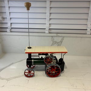 Può includere: Un modello di trattore a vapore Mamod con caldaia cilindrica verde scuro, ruote a raggi rosse e tetto color crema. Una lunga e sottile asta metallica con un manico in legno marrone si estende verticalmente dalla struttura del tetto del trattore.