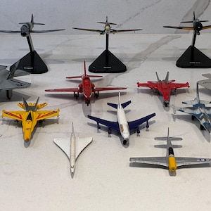 Puede incluir: Una colección de doce modelos de aviones a escala en varios colores y diseños, incluyendo aviones de combate, un avión comercial y un avión supersónico. Tres modelos están montados sobre soportes negros, mientras los otros descansan planos.