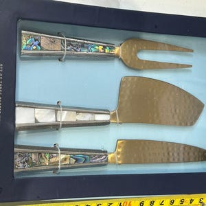 K&ouml;nnte beinhalten: Ein Set aus drei K&auml;semessern, pr&auml;sentiert in einer blauen Box. Die Messer haben goldfarbene Klingen und dekorative Griffe mit Abalone- und Perlmutt-Inlays. Auf der Box steht "Set of Three Cheese Knives" und "Spread the Love."