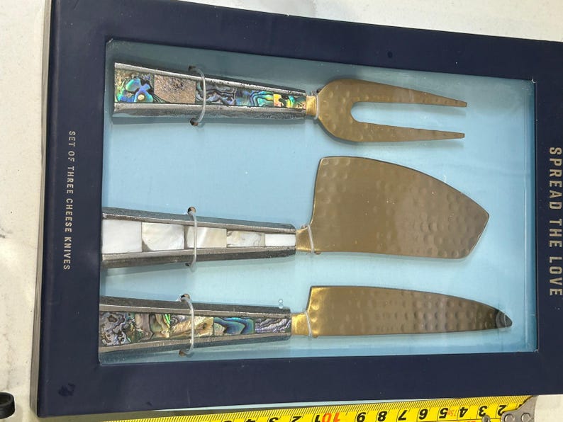 K&ouml;nnte beinhalten: Ein Set aus drei K&auml;semessern, pr&auml;sentiert in einer blauen Box. Die Messer haben goldfarbene Klingen und dekorative Griffe mit Abalone- und Perlmutt-Inlays. Auf der Box steht "Set of Three Cheese Knives" und "Spread the Love."