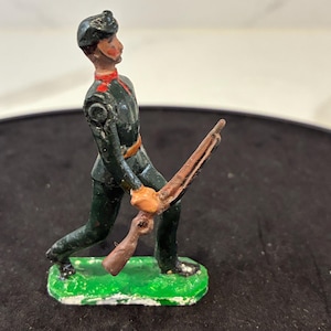 May include: A vintage painted metal toy soldier figurine standing on a green base. The figure wears a dark green uniform with red collar accents and a matching cap, holding a brown rifle in a forward-facing position with one arm extended.