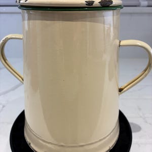 Può includere: Caffettiera in smalto color crema con coperchio e due manici. La pentola ha un bordo verde attorno al coperchio e mostra segni di usura. La pentola è appoggiata su un sottobicchiere nero.