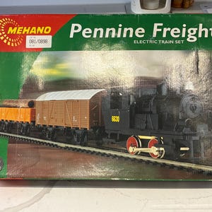 May include: A Mehano Pennine Freight electric train set box featuring a black steam locomotive with the number 6630, a brown boxcar, and an orange flatcar carrying logs on a track. The packaging is green and red with HO scale text.