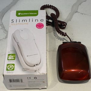 Può includere: Un telefono slimline rosso scuro lucido con cavo a spirale, posizionato accanto alla sua confezione originale in cartone bianco. La scatola riporta il testo Southern Telecom, Slimline TELEPHONE ed evidenzia funzioni come la selezione rapida e l'uso in blackout.
