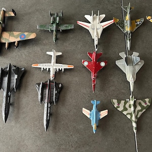 Op de afbeelding: Een verzameling van twaalf gegoten modelvliegtuigen gerangschikt in rijen. De set bevat diverse militaire vliegtuigen met camouflagepatronen, zwarte, witte, rode en blauwe kleurenschema's, met zichtbare markeringen zoals USAF en roundels.