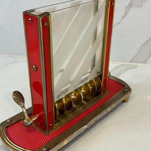 May include: A vintage cigarette dispenser featuring a rectangular clear glass case with white diagonal stripes, framed by red enamel and brass metal. The base is covered in red textured material with a brass lever and a row of brass circular holders.